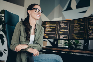 Young female programmer working at her desk in a modern workspace with dual monitors, coding and smiling confidently
