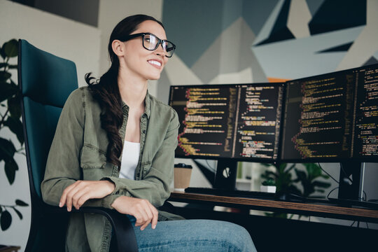 Young female programmer working at her desk in a modern workspace with dual monitors, coding and smiling confidently