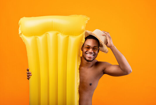 Hello summer. Positive black man in sunglasses lifting his straw hat in greeting gesture, hiding behind inflatable lilo on yellow studio background. Cheerful young guy chilling on beach