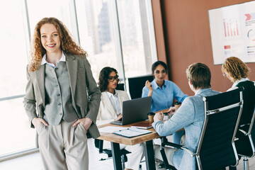 Professional businesswoman standing confidently in office setting while team collaborates on project