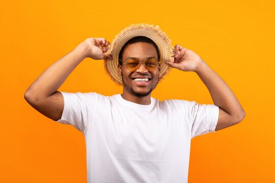 Portrait of handsome African American man in casual clothes, straw hat and sunglasses on yelow studio background. Millennial black guy wearing summer outfit, ready for beach vacation
