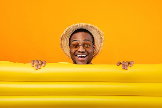 Millennial black guy in straw hat and sunglasses hiding behind inflatable lilo on yellow studio background. Young African American man with pool float having tropical summer vacation or beach party