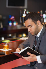 Middle Eastern man in gray suit sitting at wooden bar counter writing in notebook with drink
