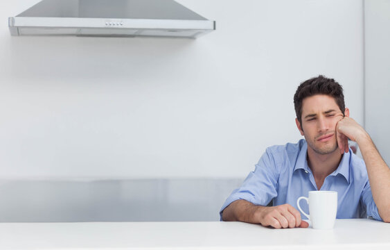 Man sitting at kitchen counter supporting head with hand while holding white mug, copy space