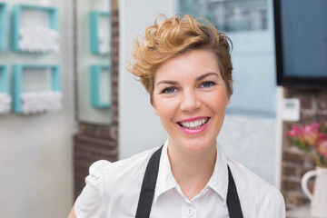 Woman barista smiling while standing behind teal shelves and menu board wearing black apron