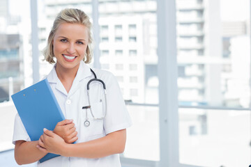 Female healthcare worker standing in clinic with clipboard, stethoscope by windows overlooking city