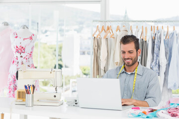 Hispanic man smiling and typing on laptop at fashion studio with sewing machine and measuring tape