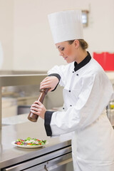 Female chef seasoning green salad with wooden pepper mill on steel countertop in clean kitchen