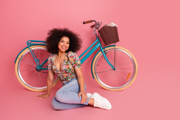 Smiling young woman with afro hairstyle sitting on a pink background beside a vintage bicycle with a basket