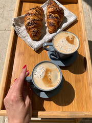 Traditional Italian breakfast. Fresh croissants and cappuccino on a wooden tray. Woman's hand holding a cup of Cappuccino, view from above. European food and pastry. Morning meal. Coffee break.