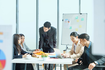 Group of Southeast Asian executives sitting around table in bright office, leader pointing at chart with confidence, representing collaboration, strategic planning, and leadership in business.