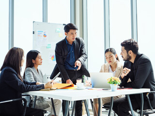 Southeast Asian business professionals in formal attire gathered in modern office, leader presenting ideas while colleagues listen attentively, symbolizing teamwork, leadership, and corporate success.