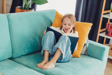 Obraz premium Happy young girl reading a book on a colorful couch in a cozy living room setting, enjoying a peaceful leisure afternoon.