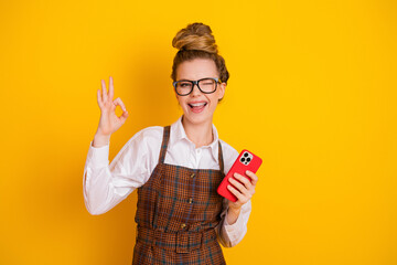 Cheerful young woman giving an OK gesture holding a smartphone against a vibrant yellow background