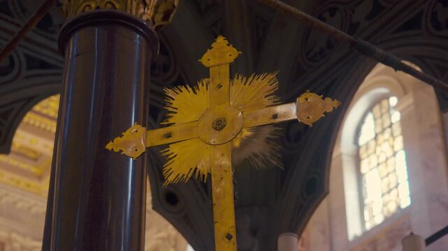 Golden processional cross featuring central sunburst detail, positioned before marble column inside historic basilica with arched window and intricate ceiling above