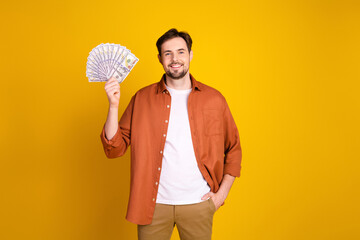 Confident young man holding money fan on yellow background, showcasing success and financial...