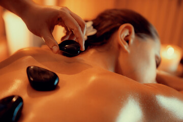 Closeup hot stone massage at spa salon in luxury resort with warm candle light, blissful woman customer enjoying spa basalt stone massage glide over body with soothing warmth. Quiescent