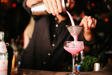A professional bartender pouring a pink cocktail from a shaker through a strainer into a glass on a...