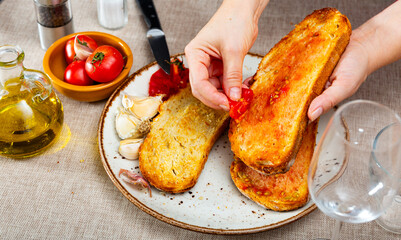 Woman preparing toast pa amb tomaquet with fresh ripe tomato, olive oil, and garlic, showcasing simplicity and flavors of traditional Mediterranean cuisine. Cropped shot..