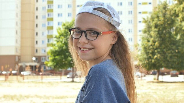 Cheerful schoolgirl wearing glasses, cap, smiling brightly near playground during sunny day - Powered by Adobe