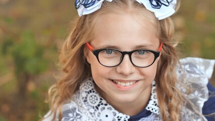 Smiling schoolgirl wearing glasses, blonde hair decorated with white and blue bows, expressing...