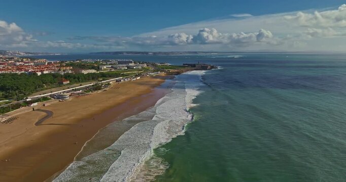 Drone view of a fortress in Portugal with the famous bridge in the background. Saint Julian Fortress with lighthouse tower from beach of Carcavelos, Portugal. Atlantic ocean waves beach in Carcavelos