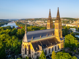 Golden morning light bathes the Vysehrad Basilica of St. Peter and St. Paul in Prague. Surrounded by greenery, the cathedral stands tall against the skyline, reflecting a tranquil moment of the dawn. © pyty
