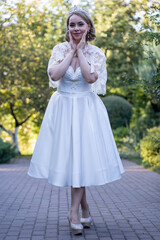 Full-body portrait of a bride in a white tea-length wedding dress, lace cape, and tiara, gently touching her cheeks with both hands in a charming and delicate gesture. Captured on a cobblestone path