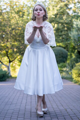 Full-body portrait of a bride in a white tea-length wedding dress and lace cape, blowing a kiss with a sweet expression. Captured outdoors on a cobblestone path surrounded by lush greenery