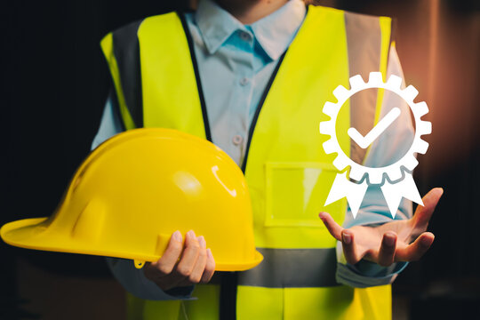 Occupational safety assurance concept. Engineer holding yellow safety helmet with quality assurance badge symbol for workplace protection compliance.