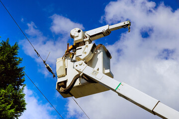 Electrical worker in bucket truck is high above, making repairs to power lines against bright sky filled with clouds.