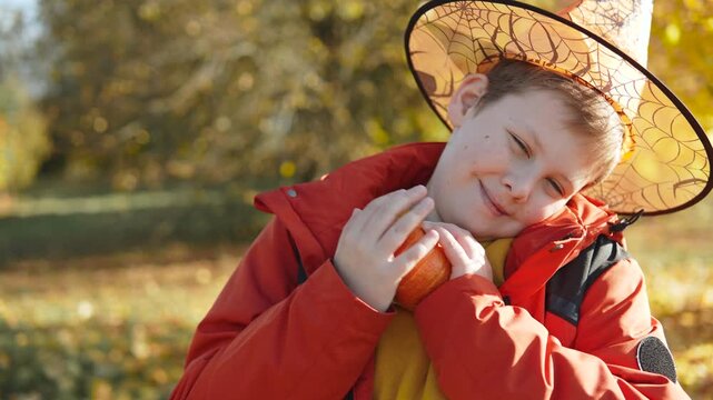 Caucasian boy in wizard cap smiles and hugs small pumpkin. Portrait of cute child with festive attribute on sunny autumn day against background of yellow trees.