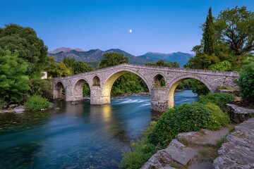 Fototapeta premium Ancient Stone Bridge Spanning a River at Dusk with Mountain Backdrop and Moon in Konitsa Greece