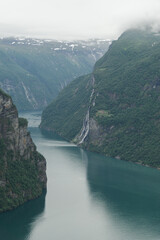 A classic view of Geirangerfjord, a Norwegian fjord with steep, green mountains and a cascading waterfall called "The Seven Sisters." The calm, dark water reflects the sky, with snow patches visible 
