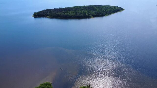 A large, green island stands in the center of a large, shimmering lake at the mouth of a river in an agricultural valley. Lake Matapedia, Quebec, Canada, 2025.