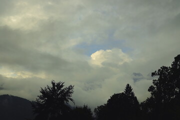 Dramatic cloudy sky with trees silhouette and mountain peak,Dark silhouette of trees against a dramatic cloudy sky with a small patch of blue and a bright white cloud in the center. Moody and atmosphe