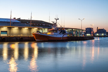 Single fishing boat moored in a calm harbor at twilight, illuminated by soft glowing lights.