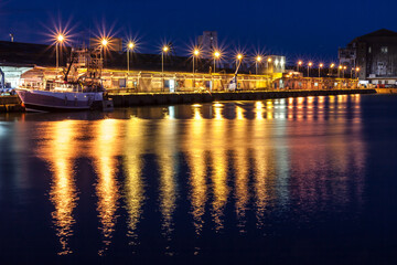Fishing harbor illuminated by bright lights at night, with golden reflections shimmering across calm waters.