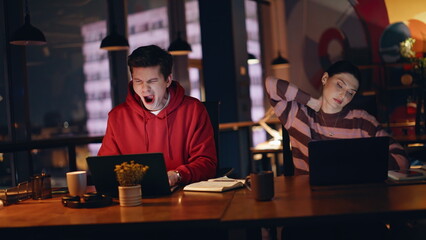 Sleepy colleagues looking laptops working overtime at dark office closeup.