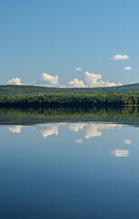 Calm lake reflecting forested hills and clouds under clear blue sky, peaceful nature landscape.