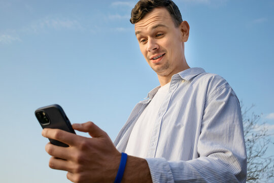 A young man in a casual shirt holding a smartphone and looking at the screen with a surprised and amused expression, standing outdoors under a clear blue sky.