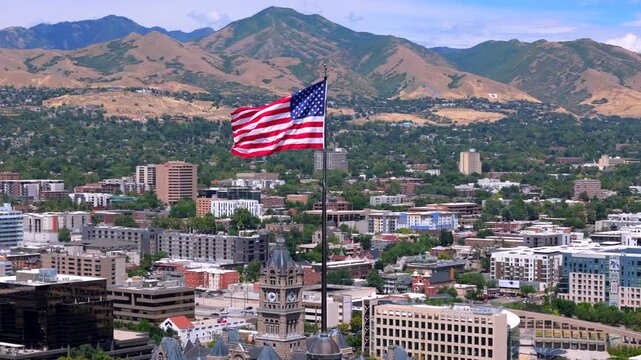 Aerial footage shows an American flag waving on a tall pole, with a vibrant Salt Lake City, Utah and majestic mountains in the background, symbolizing freedom and patriotism for all viewers