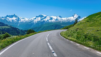 Fototapeta premium Mountain road curves through alpine meadows towards snow-capped peaks; summer travel