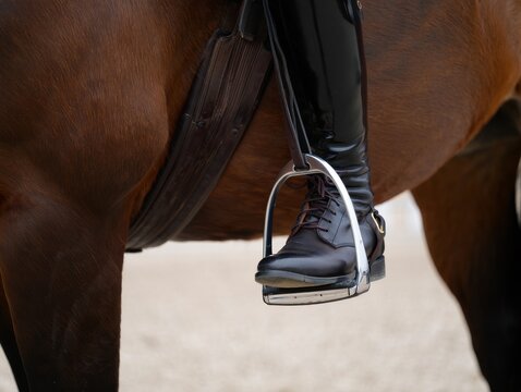 Extreme close-up of an equestrian's black leather riding boot securely placed in a metal stirrup on the side of a brown horse.
