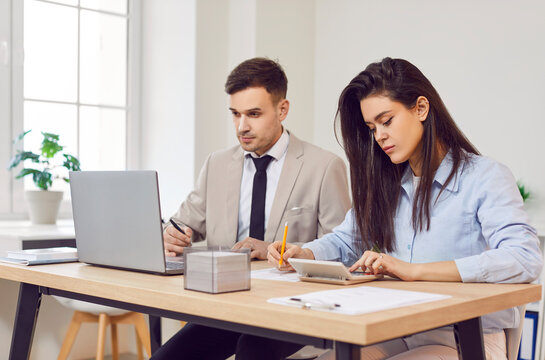 Two concentrated business people work in office, sitting together at desk. Female accountant using calculator with male colleague working on laptop, calculating monthly tax, analyzing financial data.