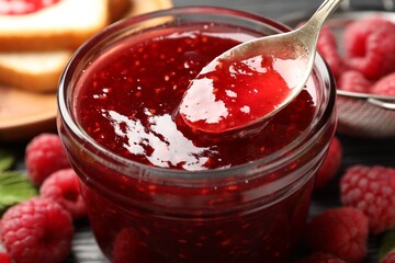 Taking sweet raspberry jam with spoon from glass jar on table, closeup