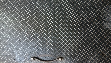 This is a close-up shot of a dark, metallic surface. The surface is textured with a repeating diamond-plate pattern, which gives it a grimy, industrial look. The metal is a dark, almost charcoal gray,