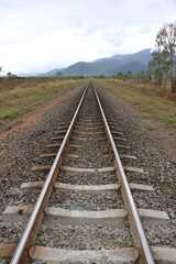 Obraz premium Railway tracks leading into the distance surrounded by gravel, grass, trees and mountains