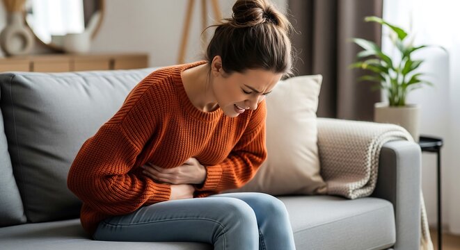 A young woman sits on a grey couch, clutching her stomach in apparent pain. She is wearing an orange sweater and jeans, exhibiting signs of discomfort or illness. Her expression is one of di