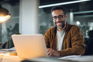 A smiling man working on a laptop in a modern office environment, with warm lighting and plants in the background, showcasing a productive and relaxed atmosphere